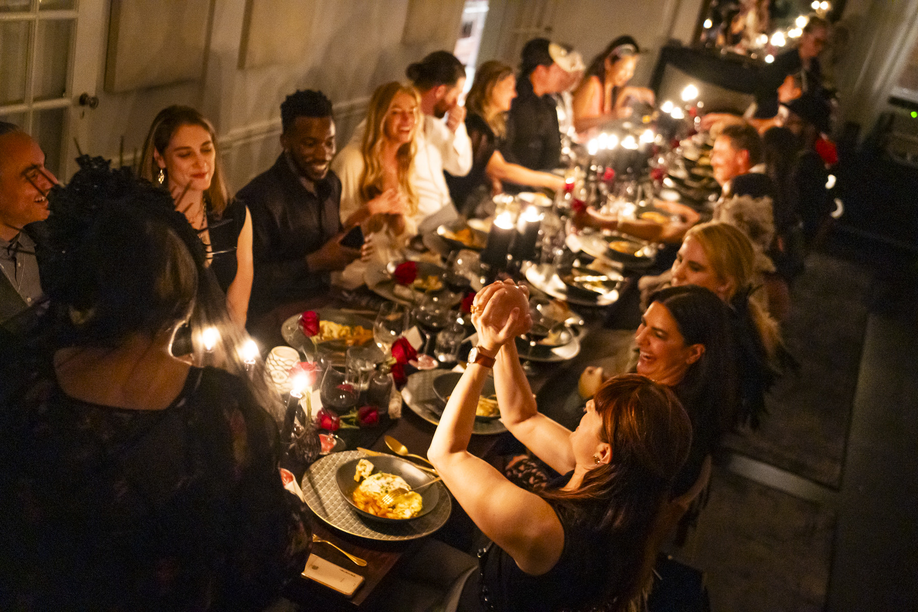 Guests laughing around a long candlelit dinner table, hands clasped mid-toast